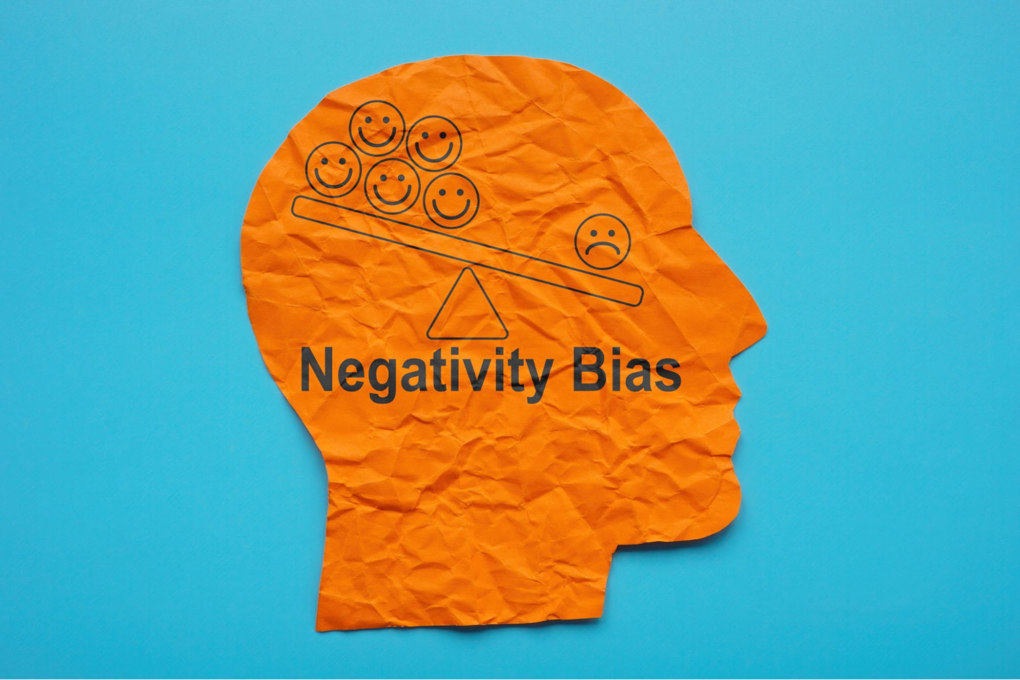 An orange paper silhouette of a head with a seesaw balancing happy and sad faces, labeled "Negativity Bias